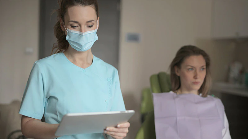 Frau, die medizinische Maske trägt, steht mit Windows 10 Tablet-Gerät, während der Patient im Zahnmedizin-Stuhl sitzt