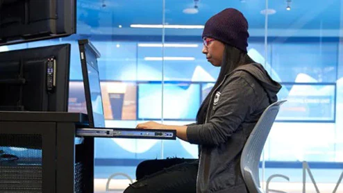 Woman in a stocking hat and hooded sweatshirt working on a network server station inside a secure room